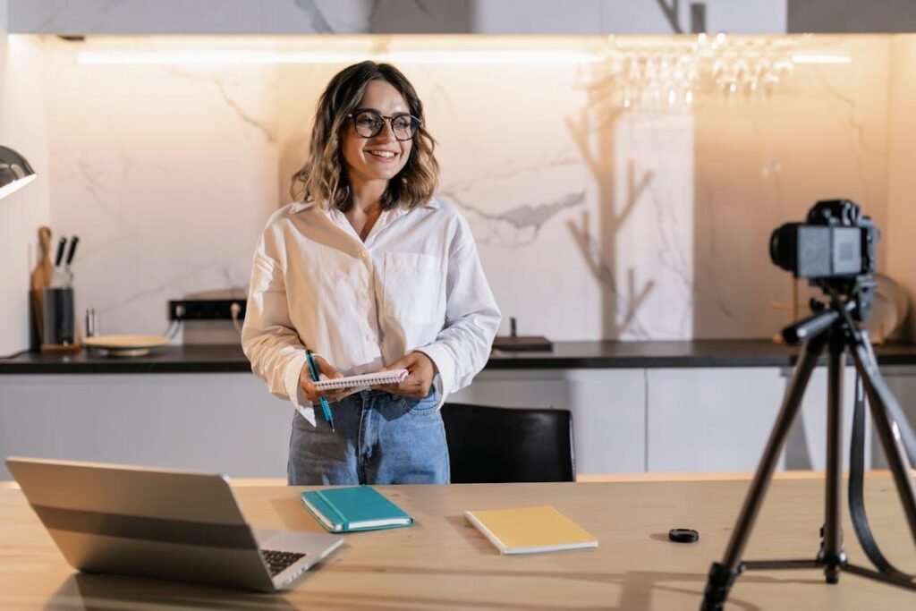 A female content creator smiling and holding a notepad while recording a video on a camera mounted on a tripod in a modern home studio setup