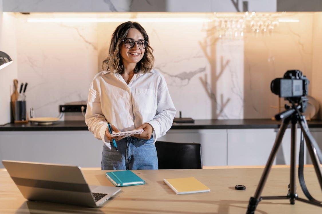 A female content creator smiling and holding a notepad while recording a video on a camera mounted on a tripod in a modern home studio setup